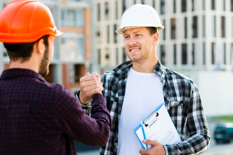 Two construction workers wearing hard hats shaking hands outdoors. One is holding a clipboard with a contract, while the other wears a red plaid shirt. Buildings under construction are visible in the background.