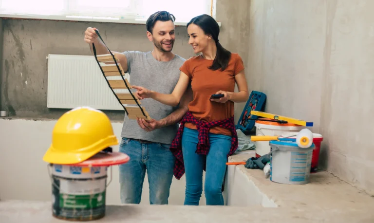 A smiling couple in a partially renovated room, discussing color samples while holding a wooden swatch. Construction tools, a yellow hard hat, paint buckets, and other renovation materials are scattered around.
