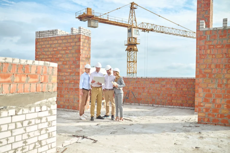A group of four engineers and architects wearing white hard hats, standing on an unfinished construction site, reviewing project plans on a laptop. A crane and partially built brick walls are visible in the background.